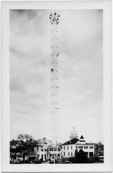 23020a Tall metal tower with person climbing, buildings and a dome in the background, cloudy sky above.
